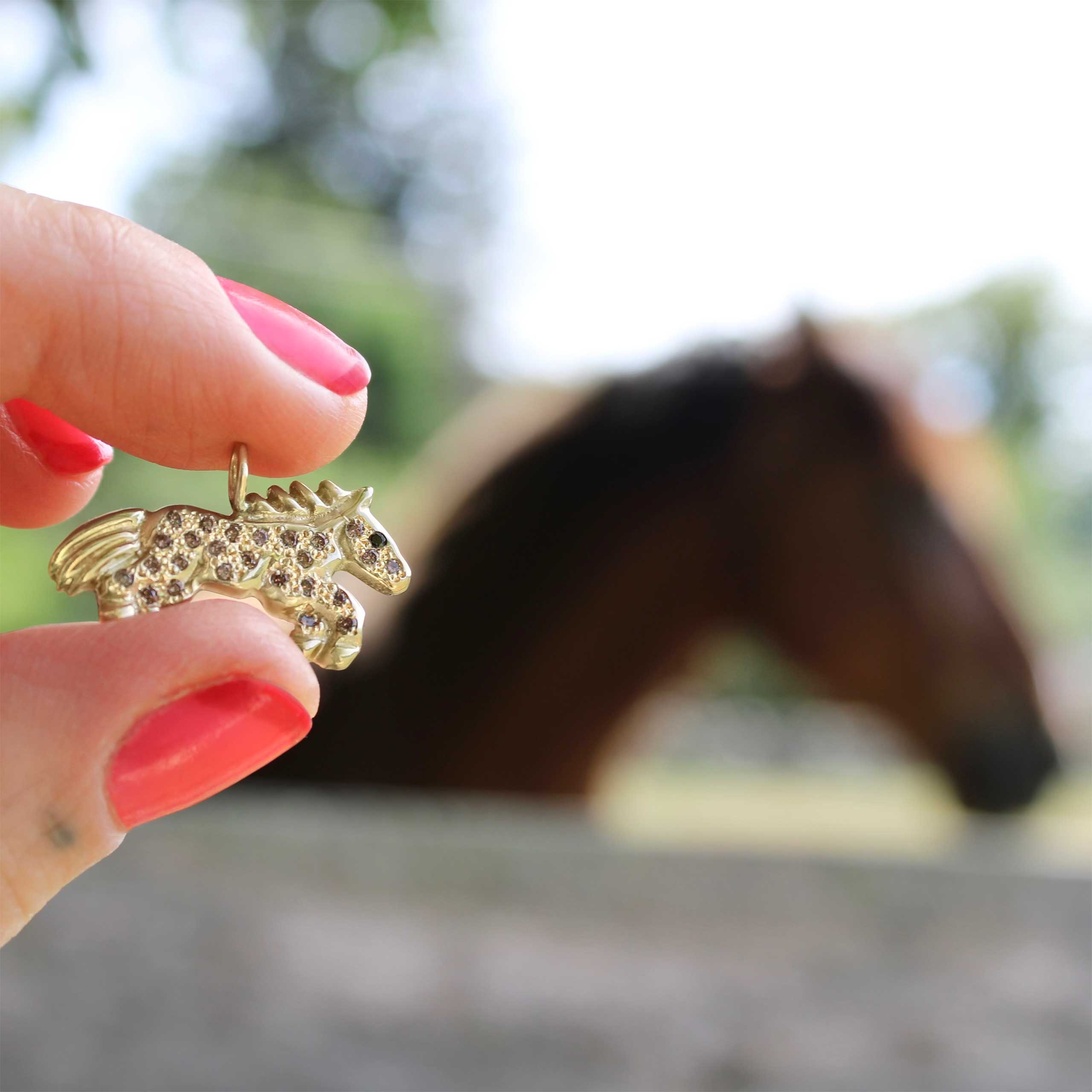 holding up a gold horse charm in the foreground with a horse in the background